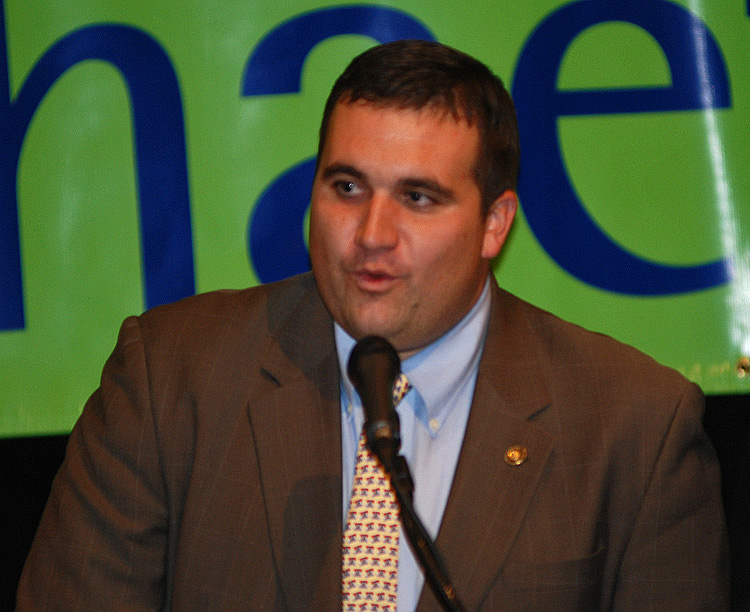 A man speaks into a microphone while wearing a suit and tie, discussing television.